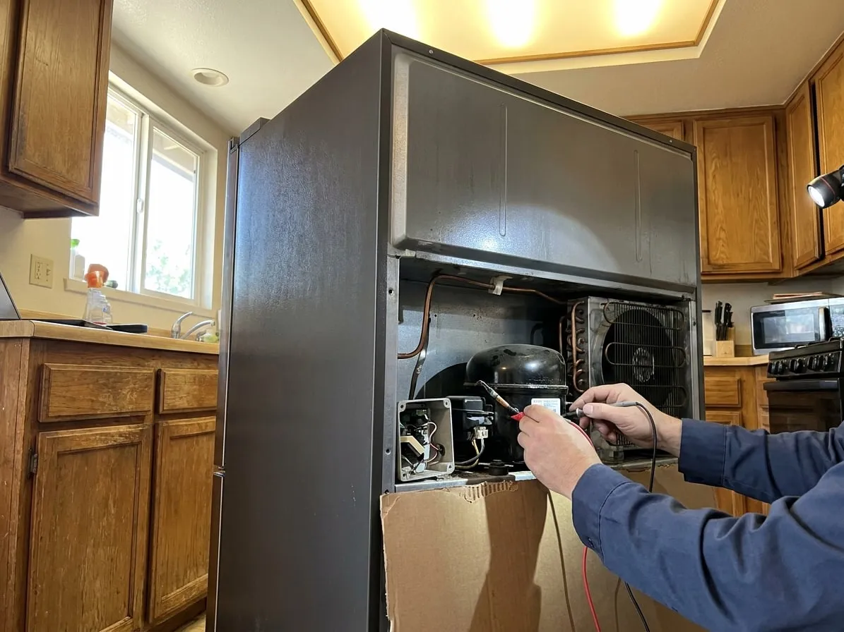 Multimeter probes on the compressor connector behind a refrigerator with the rear access cover removed